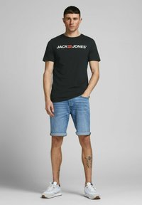 Schwarzes Baumwoll-T-Shirt mit weißem "JACK & JONES"-Logo, kombiniert mit hochgekrempelten Jeansshorts und weißen Sportschuhen. Flache, glatte Texturen.