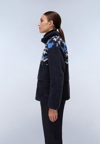 Giacca in pile blu navy con un yoke decorato in blu e bianco. Collo alto e sezione inferiore in tessuto liscio con una tasca frontale.