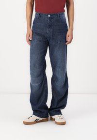 Blue striped denim pants with a relaxed fit, front pockets, and reinforced knee patches. Paired with beige sneakers.
