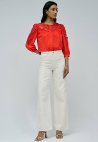 Blusa roja de botones con detalle de encaje en las mangas, combinada con jeans anchos de tiro alto blancos y sandalias de tacón dorado. Textura suave.