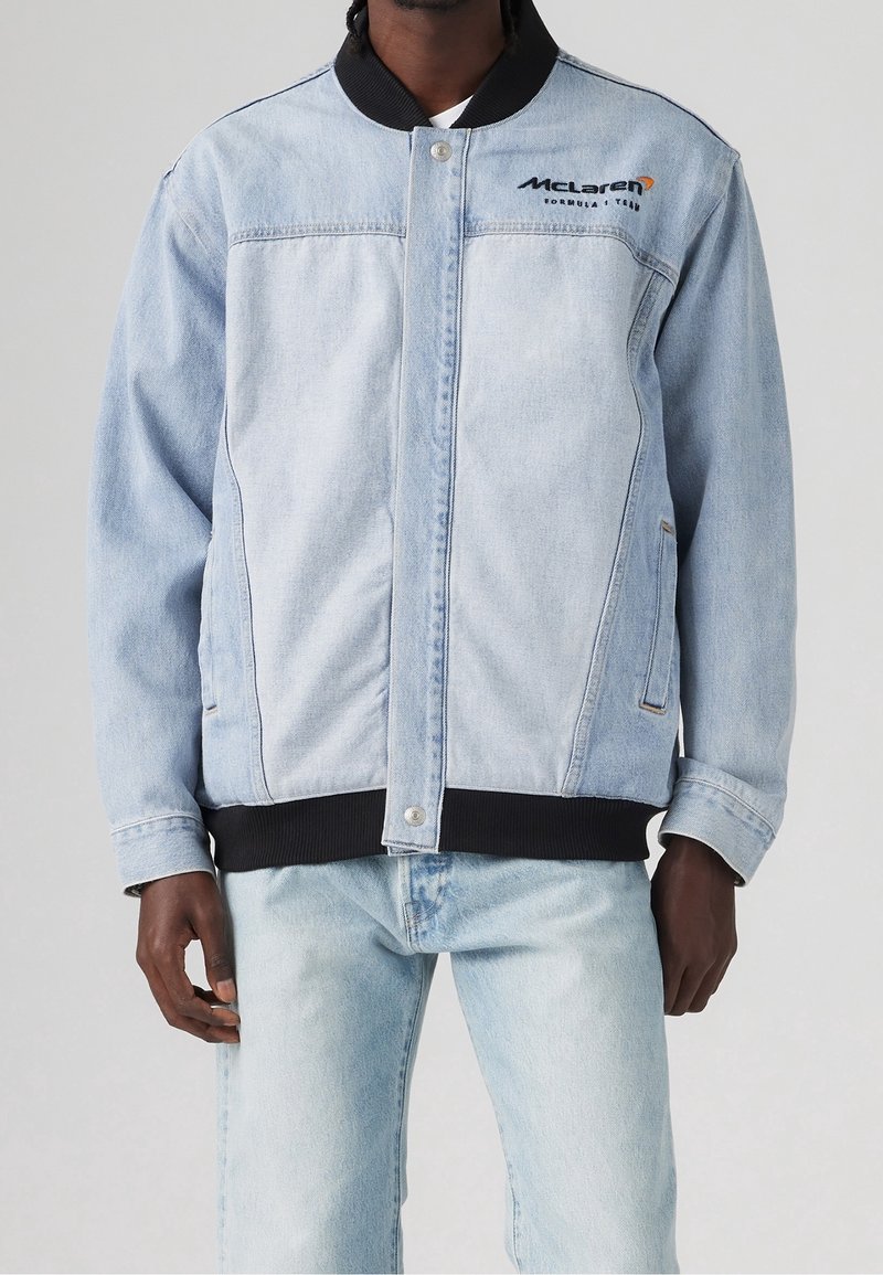 Jasnoniebieska kurtka dżinsowa z czarnym prążkowanym kołnierzem i mankietami. Na piersi znajduje się logo McLaren, dwie boczne kieszenie oraz zapięcie na zatrzaski.