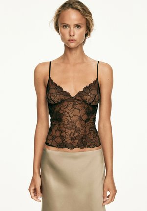 Junge Frau mit hellem Haar, die ein schwarzes Spitzen-Camisole-Top und einen beigen Rock trägt, vor einem schlichten weißen Hintergrund stehend.