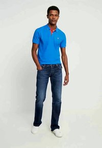 Camisa polo azul com colarinho vermelho, combinada com jeans de azul escuro. O modelo usa tênis brancos, exibindo um visual casual e ajustado.