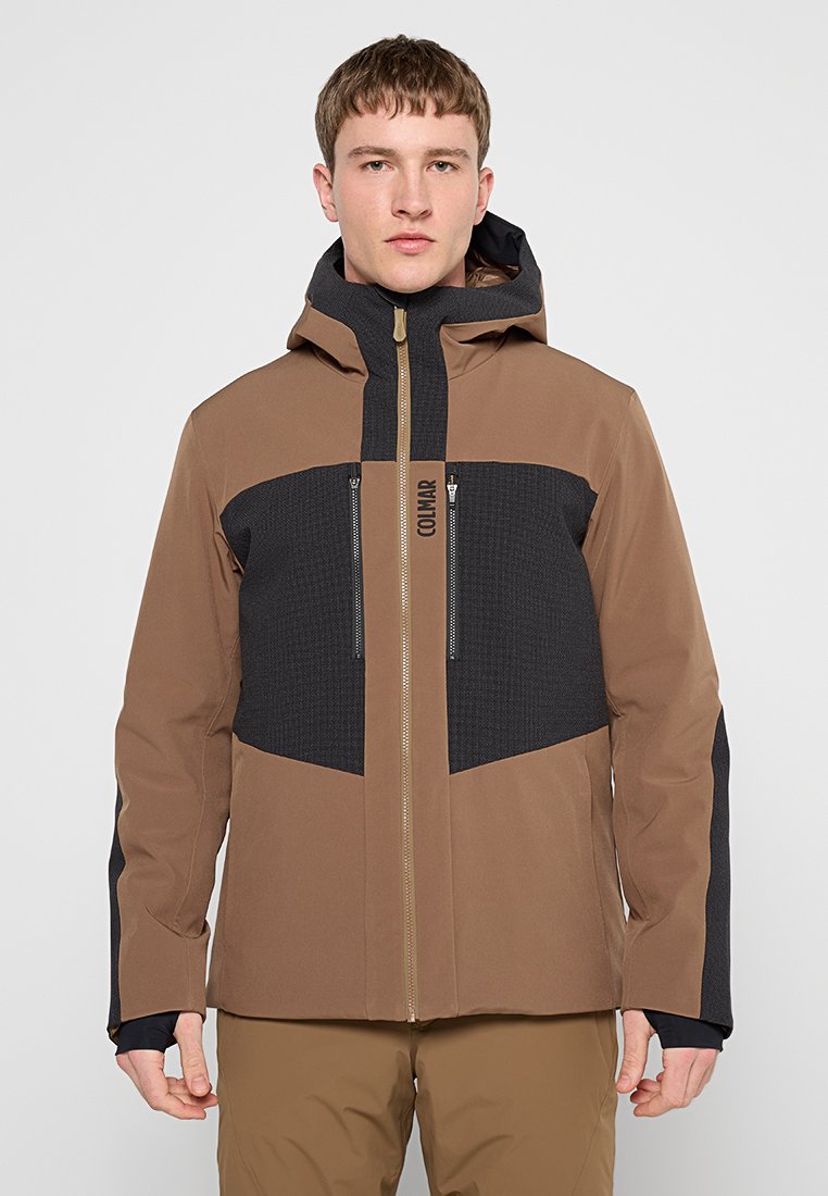 Jeune homme portant une veste à capuche marron et noire avec fermeture éclair sur le devant et poches zippées sur la poitrine, se tenant devant un fond clair.