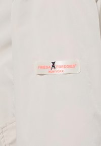 Tissu blanc avec une étiquette rectangulaire présentant une petite icône de chien noir et le texte "FRIEDA FREDDIES NEW YORK" en rose clair.