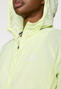 Φωτεινό κίτρινο μπουφάν με κουκούλα και κλείσιμο με φερμουάρ, με λευκό λογότυπο στο στήθος. Το υλικό φαίνεται ελαφρύ και με υφή.