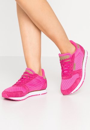 Nogi w jaskrawo różowych sneakersach z siateczki i zamszu na białych podeszwach, stojące na gładkiej, jasnoszarej podłodze.