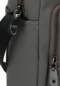Graue, strukturierte Tasche mit metallischen Reißverschlüssen und dem Markenlogo, mit glatter Oberfläche und einem seitlichen Tragegurt für zusätzlichen Tragekomfort.