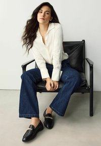 Witte knoopshirt, donkerblauwe flared jeans en zwarte loafers met een metalen accent, zittend op een zwarte stoel tegen een lichte achtergrond.
