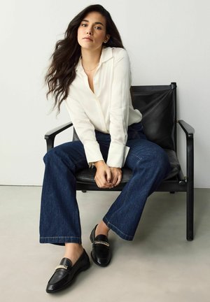 Witte knoopshirt, donkerblauwe flared jeans en zwarte loafers met een metalen accent, zittend op een zwarte stoel tegen een lichte achtergrond.