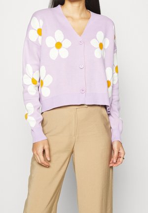Femme portant un cardigan lavande boutonné avec des motifs de marguerites blanches et jaunes et un pantalon beige sur un fond uni.