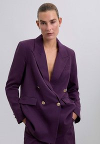 Blazer morado de doble botonadura con solapas afiladas, botones dorados y dos bolsillos frontales. Textura suave con un escote pronunciado.