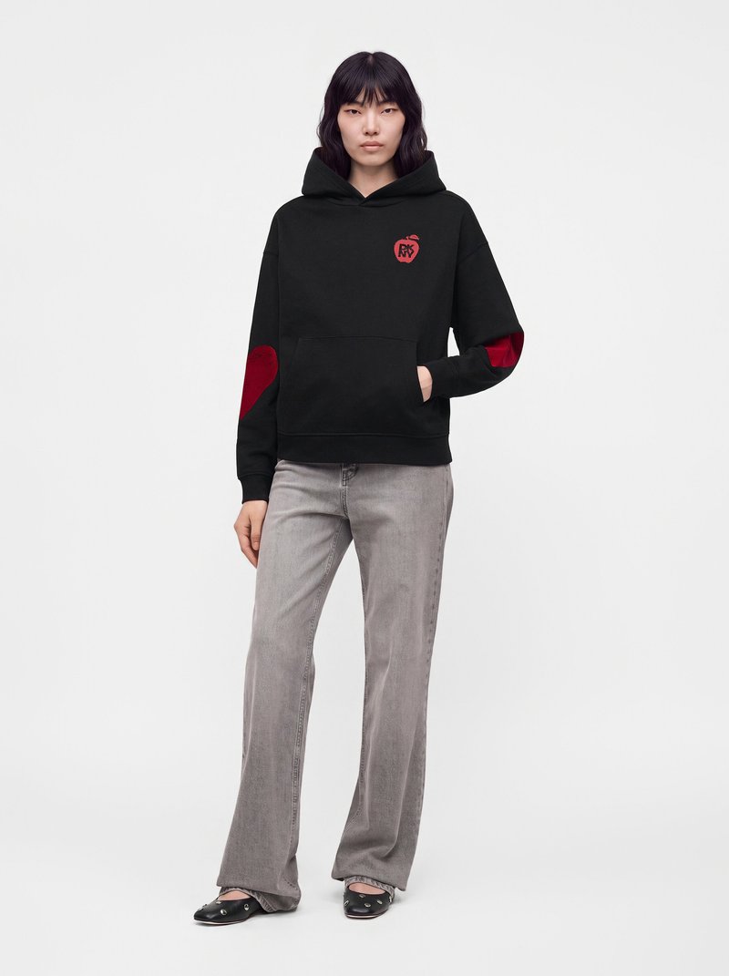 Femme portant un sweat à capuche noir avec des patchs en forme de cœur rouge sur les manches et la poitrine, un jean gris et des ballerines noires, debout devant un fond uni.