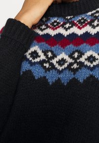 Gros plan d'une personne portant un pull en maille noire avec un empiècement à motif aux couleurs bleu, blanc, rouge et noir, aux dessins géométriques.