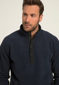 Pullover in pile della marina con una patta nera a bottoni a pressione e colletto alto. Caratterizzato da un piccolo logo sul petto e una texture morbida.