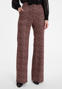 Vrouw in hoge taille wijde broek met een bruin en roze geometrisch patroon, zwarte sheer blouse en zwarte puntige schoenen.