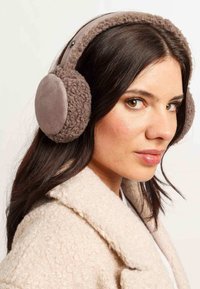 Donna con lunghi capelli scuri che indossa cuffie di peluche marroni e un cappotto beige, che guarda leggermente di lato.
