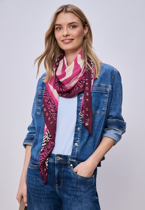 Uśmiechnięta kobieta o blond włosach, ubrana w niebieską dżinsową kurtkę i dżinsy, jasnoniebieski top oraz różowy szalik w wzory wokół szyi.