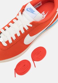 Red canvas sneaker with white accents, featuring a blue logo. Includes two pairs of red shoelaces. Rubber sole with textured details.