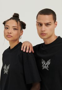 Černé bavlněné trička mají středový design stříbrného motýla v plamenech. Krátké rukávy a volný střih vytvářejí pohodlný siluetu.