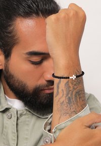 Perlenarmband mit schwarzen Perlen und einem silbernen Kreuzakzent, getragen an einem Handgelenk mit sichtbarem Tattoo, vor einem neutralen Hintergrund.