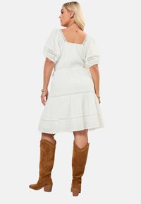 Vestido de lino blanco con mangas cortas abullonadas, cuerpo fruncido y dobladillo en capas. Combinado con botas de ante marrón hasta la rodilla con costuras decorativas.