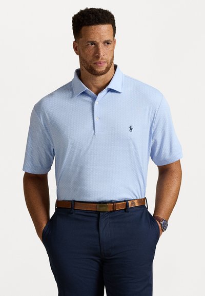 Man in een lichtblauw poloshirt, marineblauwe broek, een leren bruine riem en een horloge, met zijn handen in zijn zakken, kijkt iets opzij.