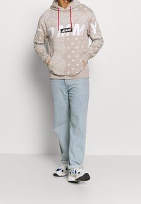 Felpa beige con motivi paisley e pois, lacci rossi e un grande logo bianco "TOMMY JEANS". Indossata con jeans azzurri e sneakers.