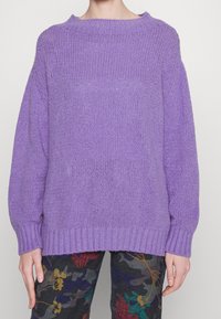 Personne portant un pull lavande à mailles lâches avec un col, des poignets et un ourlet côtelés, associé à un pantalon noir à motif floral.