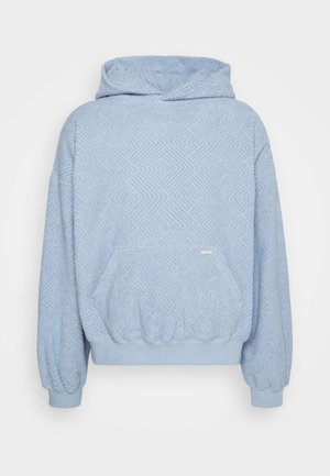 Person trägt einen blauen, strukturierten Hoodie, schwarze Hosen und schwarze High-Top-Stiefel und sitzt auf einem silbernen Metall-Aufbewahrungskoffer auf blauem Boden.
