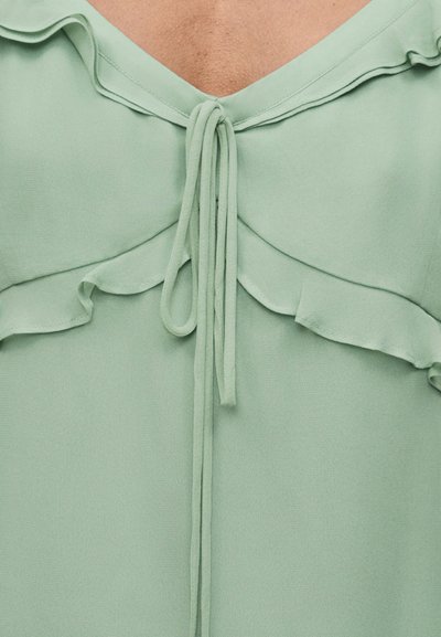 Κοντινό πλάνο ανοιχτοπράσινου μπλουζιού με βολάν στον λαιμό και δεμένα κορδόνια μπροστά στο πάνω μέρος του στήθους ενός ατόμου.