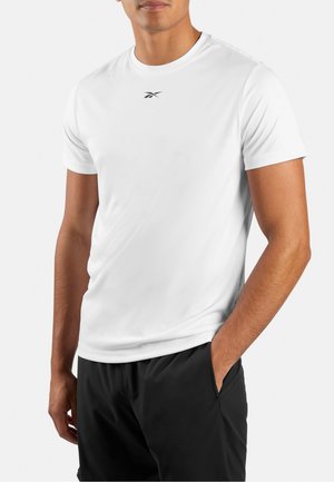 Man met een effen wit T-shirt met korte mouwen met een klein zwart logo op de borst en zwarte broek, één hand in de zak.