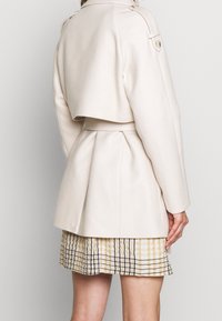 Jasno beżowa kurtka z krótkim tyłem, paskiem w talii i wyraźnymi ramionami. Posiada akcenty w postaci guzików oraz gładką fakturę.