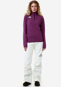Maglietta lunga viola con mezzo zip e dettaglio del logo, abbinata a pantaloni bianchi con logo nero. Entrambi gli articoli hanno un design aderente.