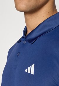 Námořnická modrá polo košile s hladkým povrchem, dvěma knoflíky a bílým logem, které obsahuje tři vzestupné pruhy na levé straně.