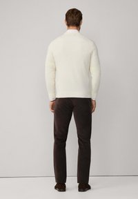 Cremfarbener Pullover mit langen Ärmeln, gerippten Bündchen und Saum, kombiniert mit braunen Hosen. Glatte Textur und taillierte Form, von hinten betrachtet.