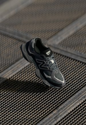 Czarno-szary but sportowy New Balance unoszący się nad teksturowaną metalową powierzchnią siatki, ze cieniem poniżej podkreślającym jego kształt.