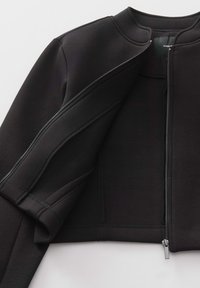 Veste noire courte en tissu doux, avec un col rond, une fermeture éclair apparente et des manches à poignet avec des coutures épurées.
