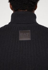 Čierny pletený sveter s vysokým golierom a štvorcovou koženou nášivkou na chrbte, s embosovaným nápisom "EA7 Emporio Armani."