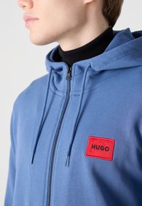 Blå hettegenser med glidelås laget av mykt stoff, med en frontlomme, snorer og en kontrasterende rød logo-patch på brystet.