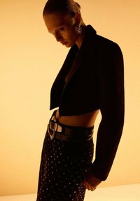 Mujer con el pelo recogido hacia atrás, usando grandes aros, una chaqueta negra corta, pantalones negros con tachuelas y un cinturón ancho, contra una luz cálida.