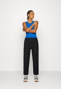 Top azul sem mangas combinado com calças pretas. Modelo a usar ténis pretos e brancos. Tecido suave e silhueta ajustada com um design casual.