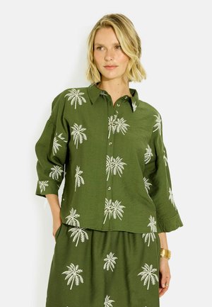 Mujer rubia con camisa suelta verde oliva y falda con estampados blancos de palmeras, mirando de lado sobre un fondo blanco.