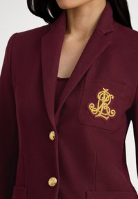 Bordo krāsas žakete ar teksturētu audumu, zelta pogām un izšūtu logo uz kabatas. Piegriezums ir pielāgots ar izgrieztiem atlokiem.