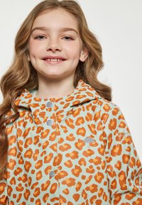 Vattentät regnjacka med en ljusblå bas och orange leopardmönster, huva, tryckknappar och en liten grå paraplygrafik.
