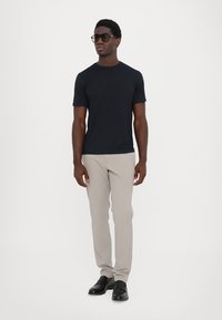 Dunkelblaues kurzärmliges T-Shirt, beige Slim-Fit-Hosen, schwarze Schuhe, Sonnenbrille. Mann steht aufrecht mit neutralem Ausdruck vor einem weißen Hintergrund.