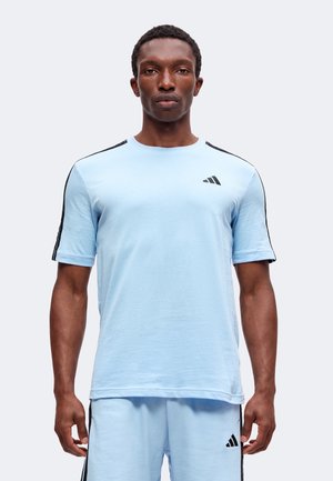 Άνδρας που φοράει αθλητική ένδυση Adidas ανοιχτού μπλε χρώματος με μαύρες ρίγες στα μανίκια και στα σορτς, στέκεται μπροστά σε απλό φωτεινό φόντο.