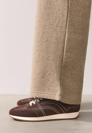 Brown suede sneakers with white laces and stitching, paired with beige, textured sweatpants that have a fitted cuff at the ankle.