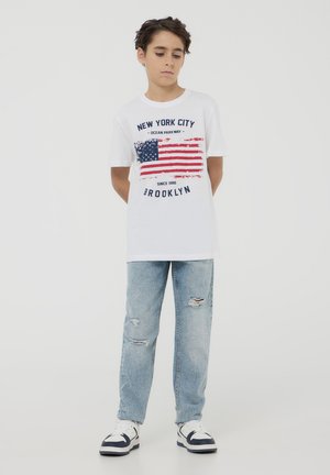 T-shirt bianco in cotone con una grafica della bandiera americana usurata e il testo "New York City", indossato con jeans strappati azzurri chiari e sneakers.
