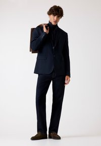 Marineblazer met twee knopen, aangevuld met een zwart top met hoge hals, donkere jeans en bruine schoenen. Het model houdt een leren bruine tas vast.